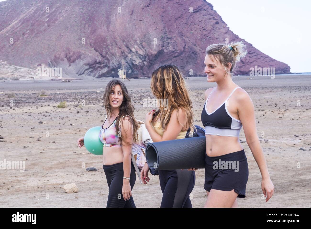 Three female friends in sports clothing with exercise equipments walking on beach. Young women using yoga mat and ball for workout at beach during vac Stock Photo
