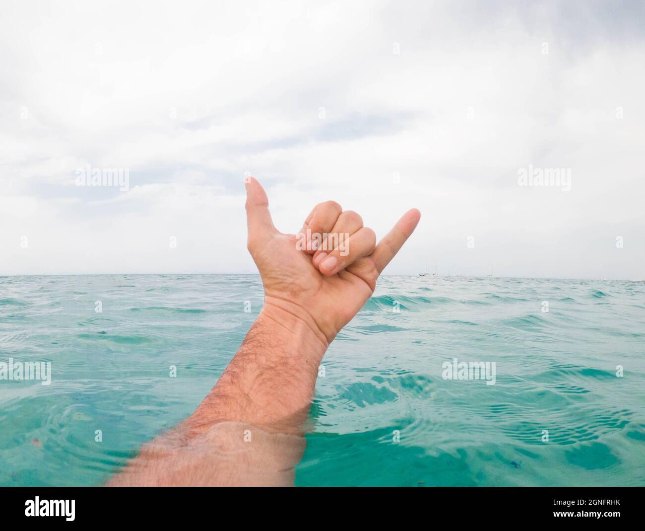 Close up of man's hand showing shaka sign above sea water against sky. Hand of a man coming out of sea water surface and gesturing shaka sign against Stock Photo