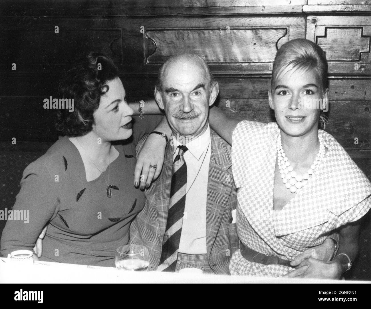 A smartly dressed elderly gentleman in a suit and tie poses for a photograph with his wife and daughter at The Society Restaurant, Jermyn Street, London. He holds them both close with his hands around their waists. Early 1960s. Stock Photo
