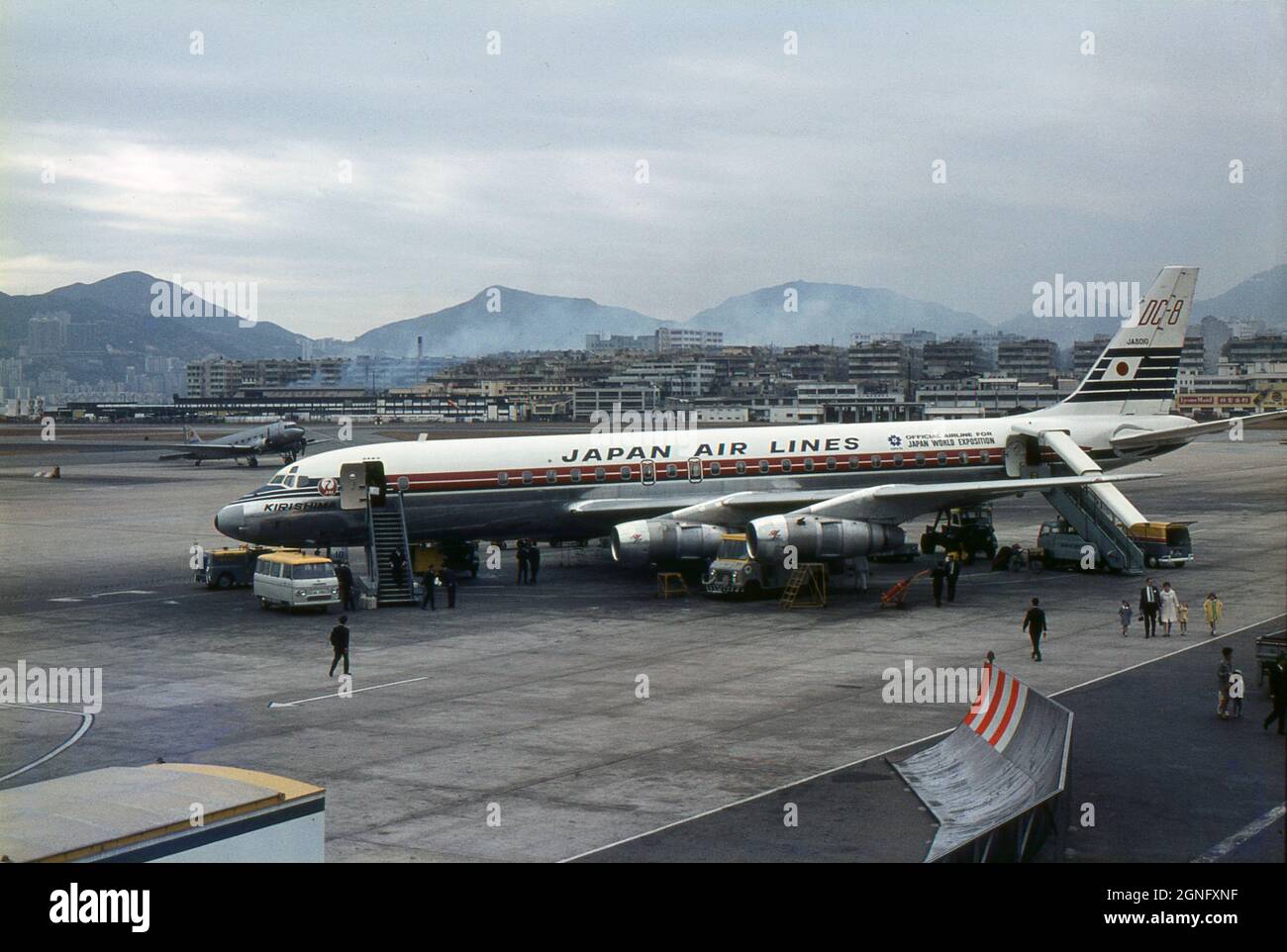 Japan Air Lines Douglas DC-8 Airliner ‘Kirishima’(JA8010) being made ready for flight at Kai Tak Airport, Hong Kong in 1968. The aircraft has the advertising slogan, “Official Airline for Japan World Exposition” painted on its fuselage, promoting the forthcoming ‘Expo '70’, the world's fair held in Suita, Osaka, Japan in 1970. Passengers are walking towards the terminal building, having disembarked from the aircraft. An ageing Douglas DC-3 propeller-driven airliner is parked in the distance. Stock Photo