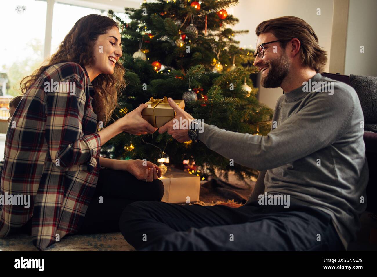 Man giving gift to his wife at home. Couple sitting by Christmas tree exchanging presents and smiling. Stock Photo
