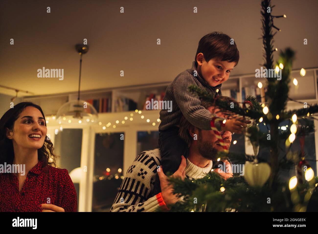 Boy sitting on his father's shoulder while decorating Christmas tree. Family decorating Christmas tree together at home. Stock Photo