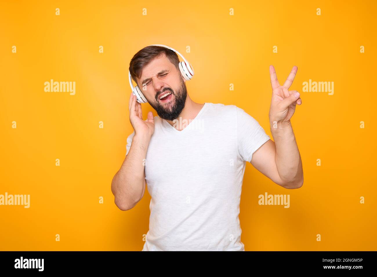 a man in white headphones with a beard, listens to music and shows the gesture of victory. Stock Photo