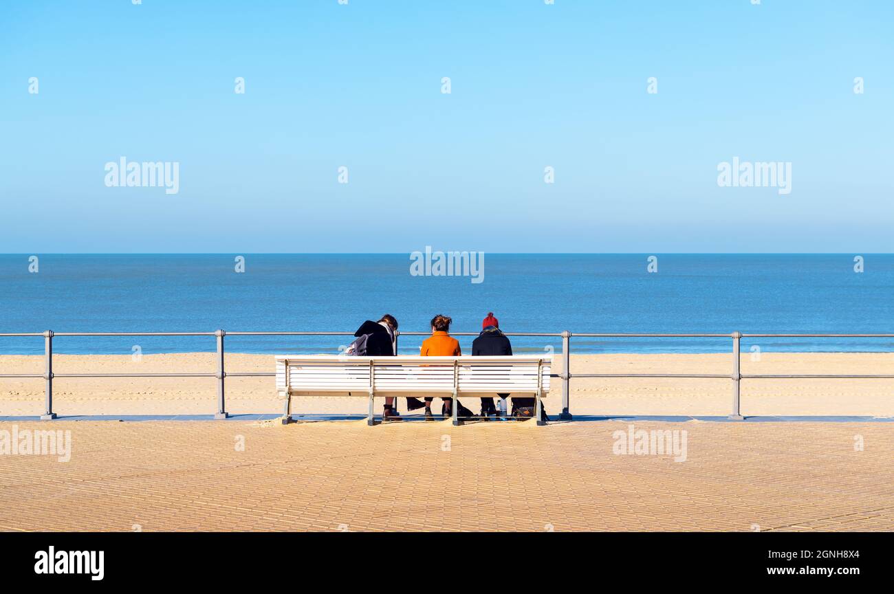 Waterfront promenade bench with three people sitting in front of Oostende (Ostend) North Sea beach, West Flanders, Belgium. Stock Photo