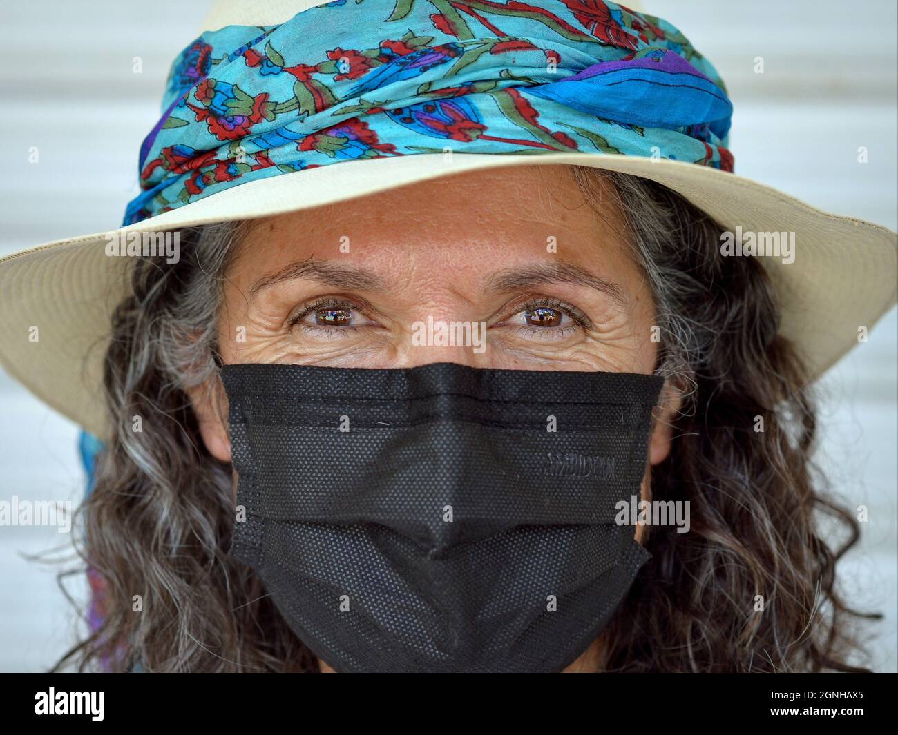Charming positive mature Caucasian woman with beautiful brown eyes wears panama hat with stylish blue hat band and black disposable face mask. Stock Photo