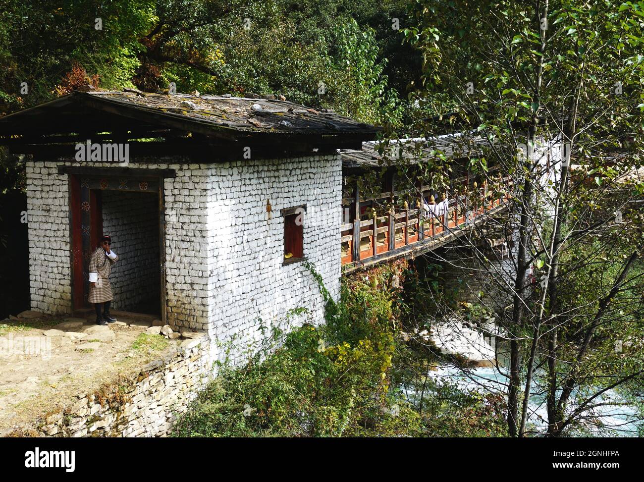 A man wearing traditional Bhutanese clothing stands at the entrance to an ancient cantilever bridge with colorful painted woodwork in rural Bhutan Stock Photo