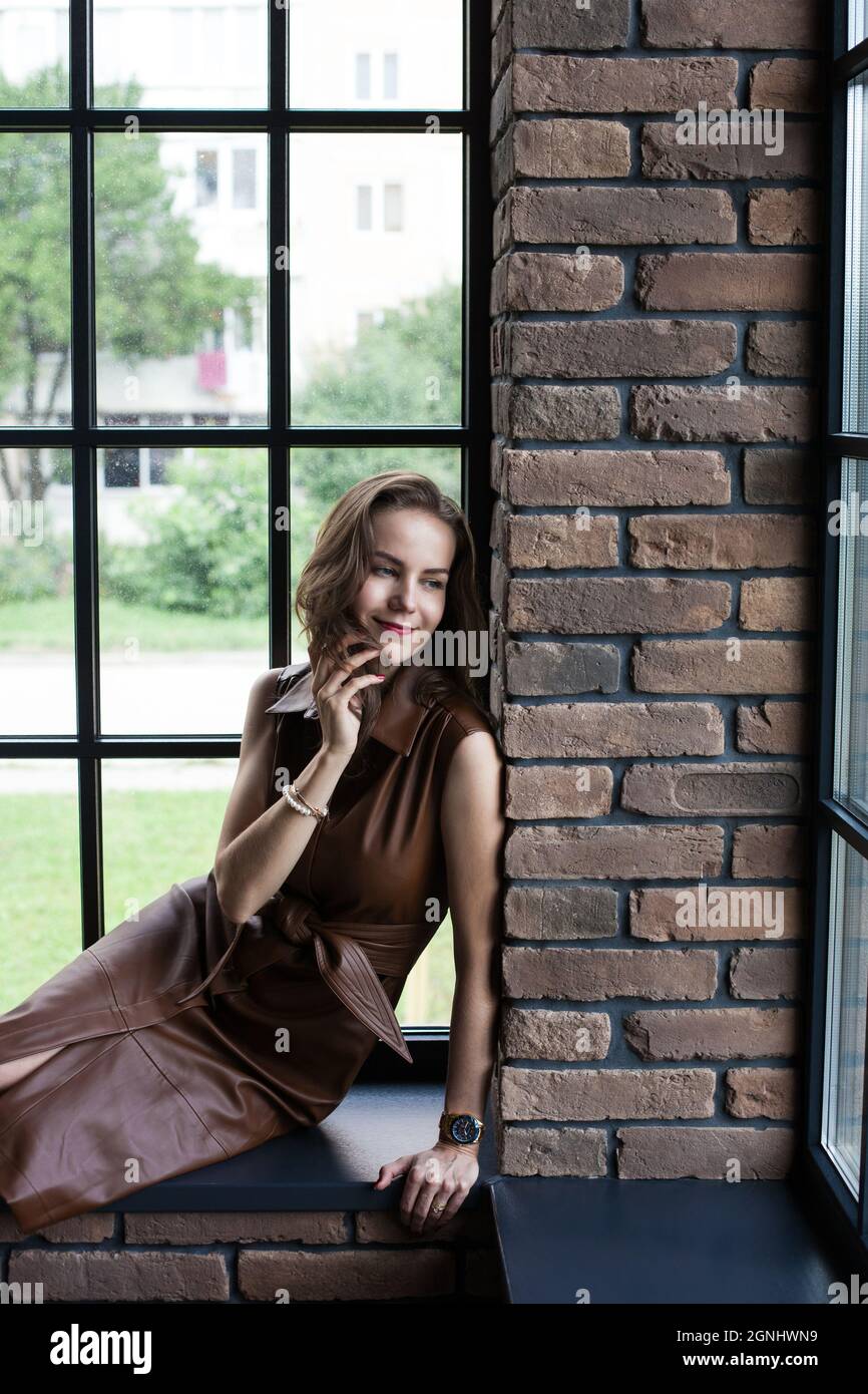 Beautiful young woman in brown leather dress sits on the windowsill ...