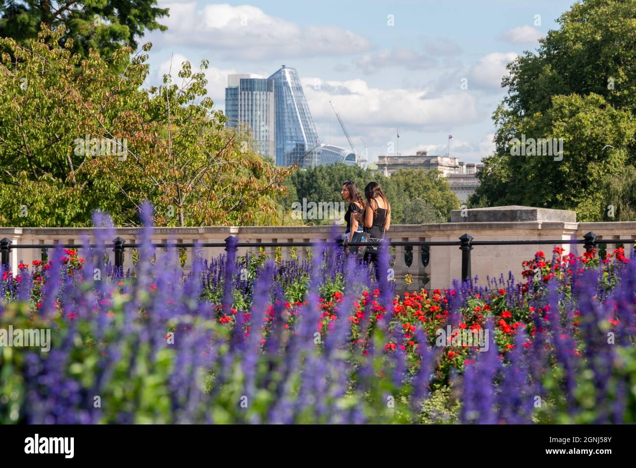 Colourful plants in the flower beds of Buckingham Palace Memorial Gardens, London, UK, with females walking past and high rise city financial district Stock Photo