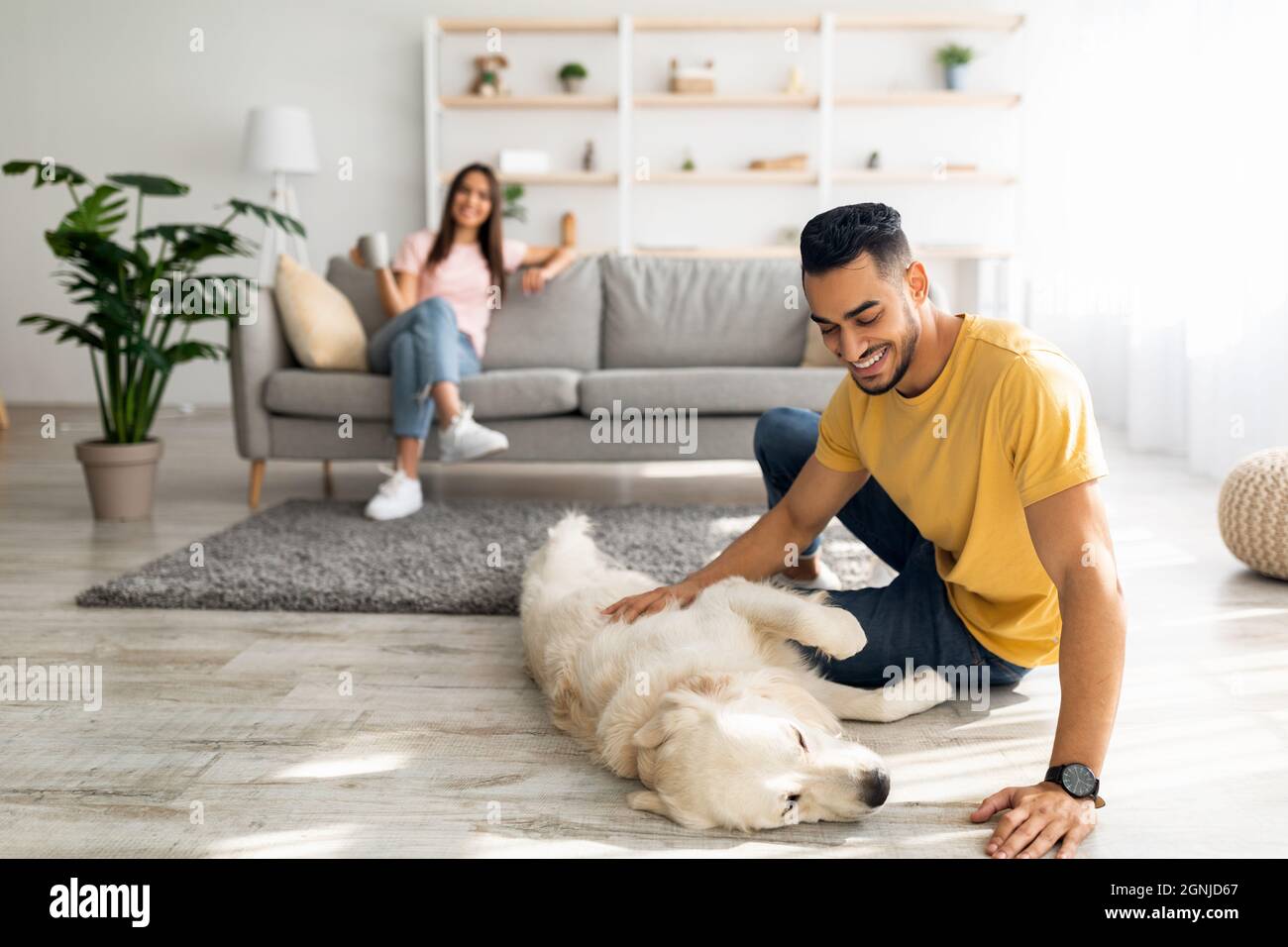 Happy Arab guy stroking cute dog on floor at home, his girlfriend relaxing with cup of coffee on background, copy space Stock Photo