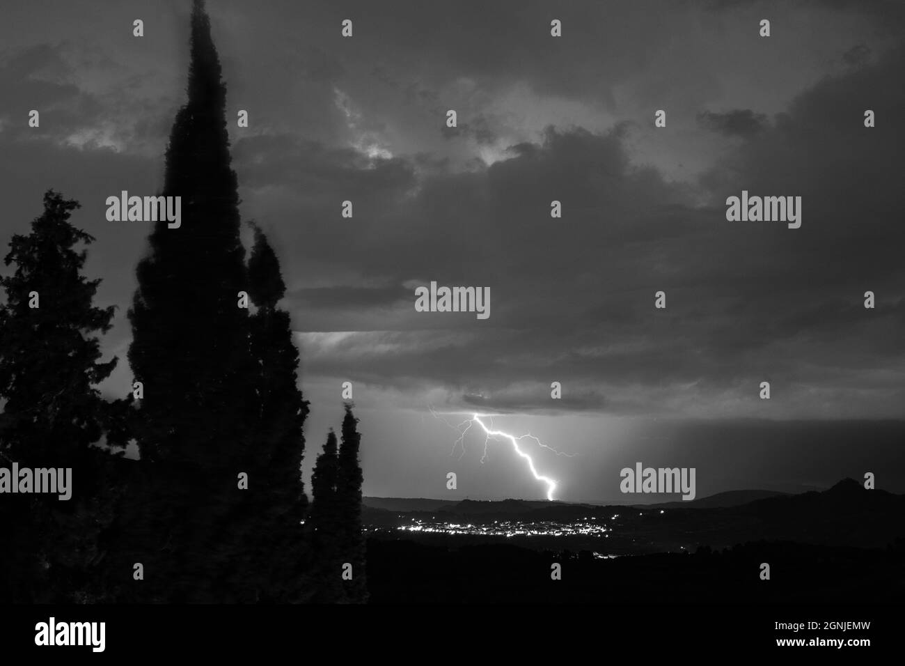 A big thunder storm passing over a town of La Llosa de Ranes with a thunderbolt touching the ground and few tree silohouettes in the foreground Stock Photo