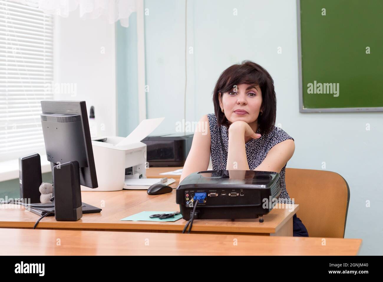 The teacher is sitting alone at her desk in the school classroom