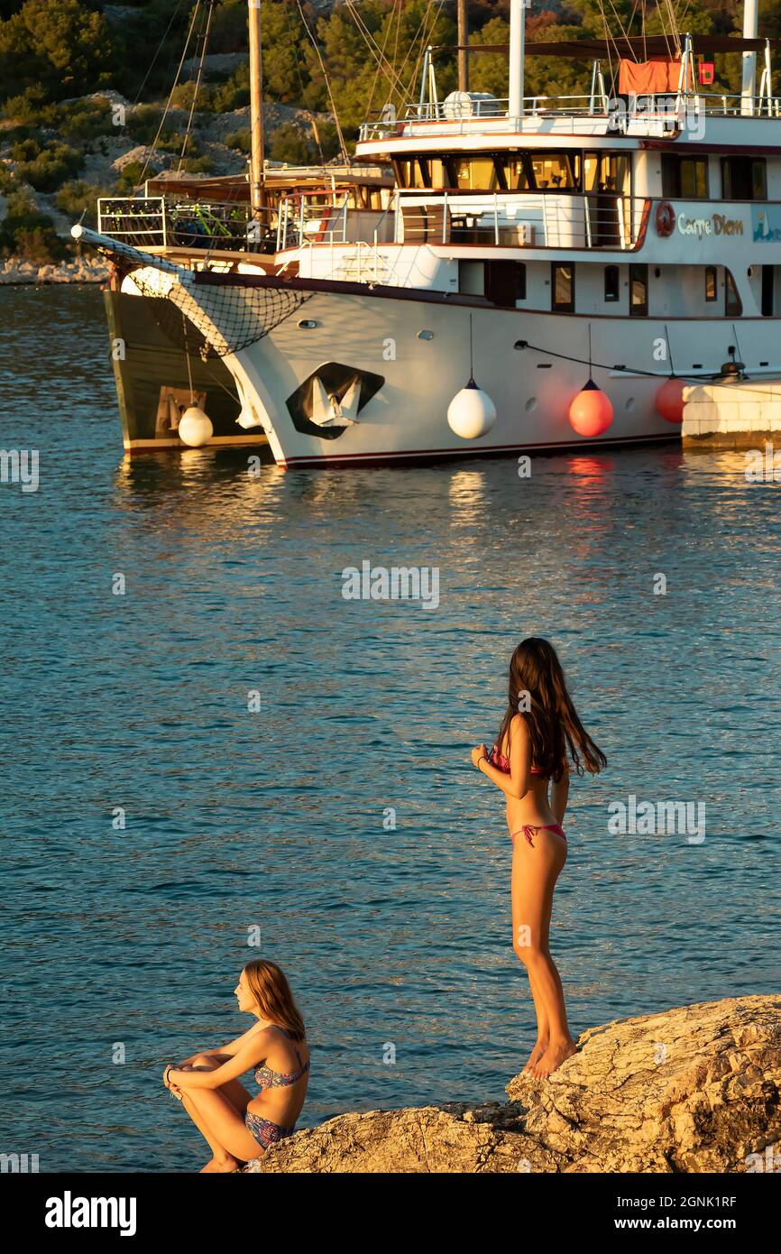 Podvrske, Murter, Croatia - August 26, 2021: Teenage girls in swimsuits standing on a golden rock, watching, and ship moored behind them, in summer su Stock Photo