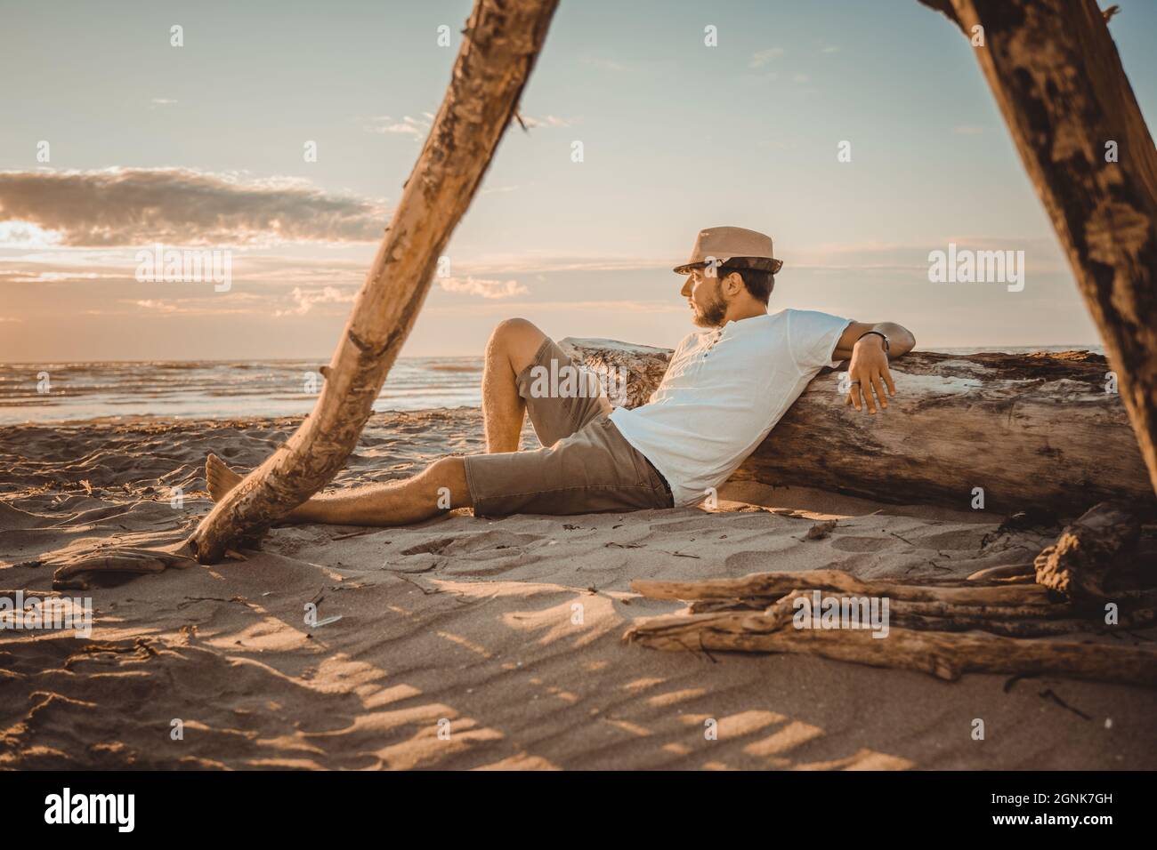 Young man relaxing with summer hat lying on the wooden stump on the desert beach with a beer  looking sunset - Close up portrait Stock Photo