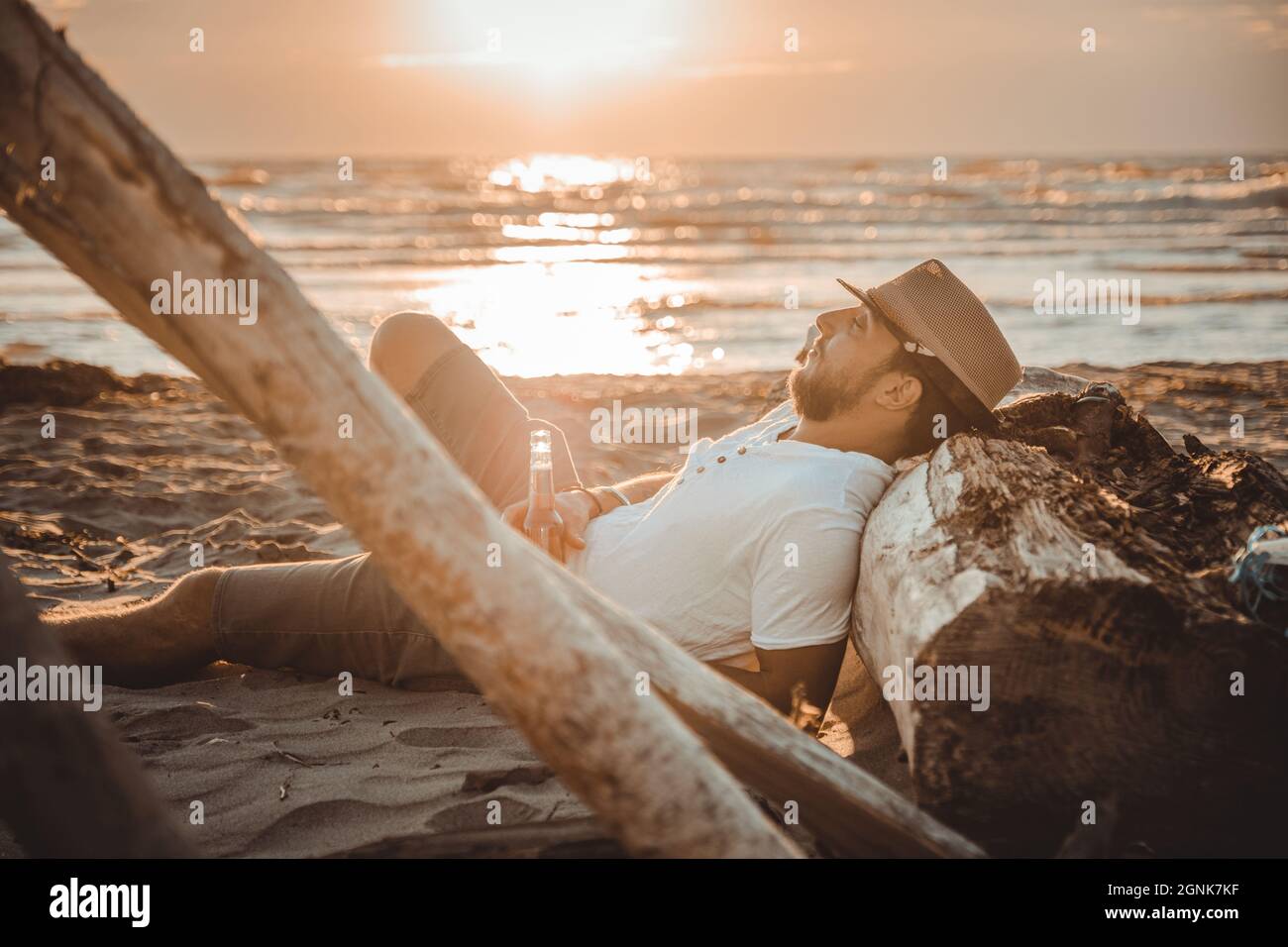 Young man relaxing with summer hat lying on the wooden stump on the desert beach with a beer  looking sunset - Close up portrait Stock Photo