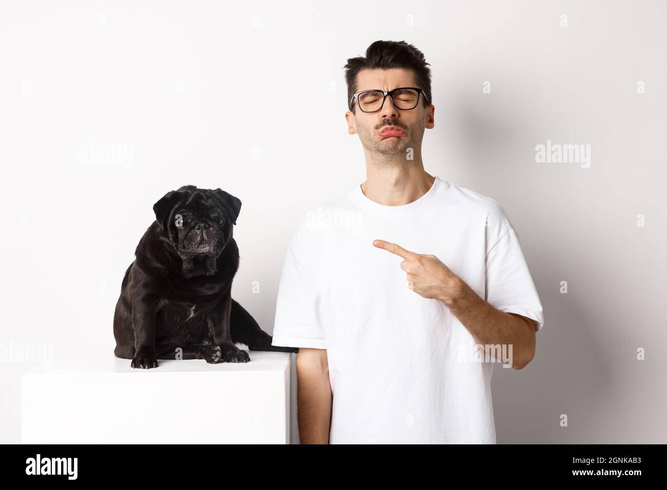 Sad and gloomy pet owner pointing at his black pug dog and sobbing, standing upster against white background Stock Photo