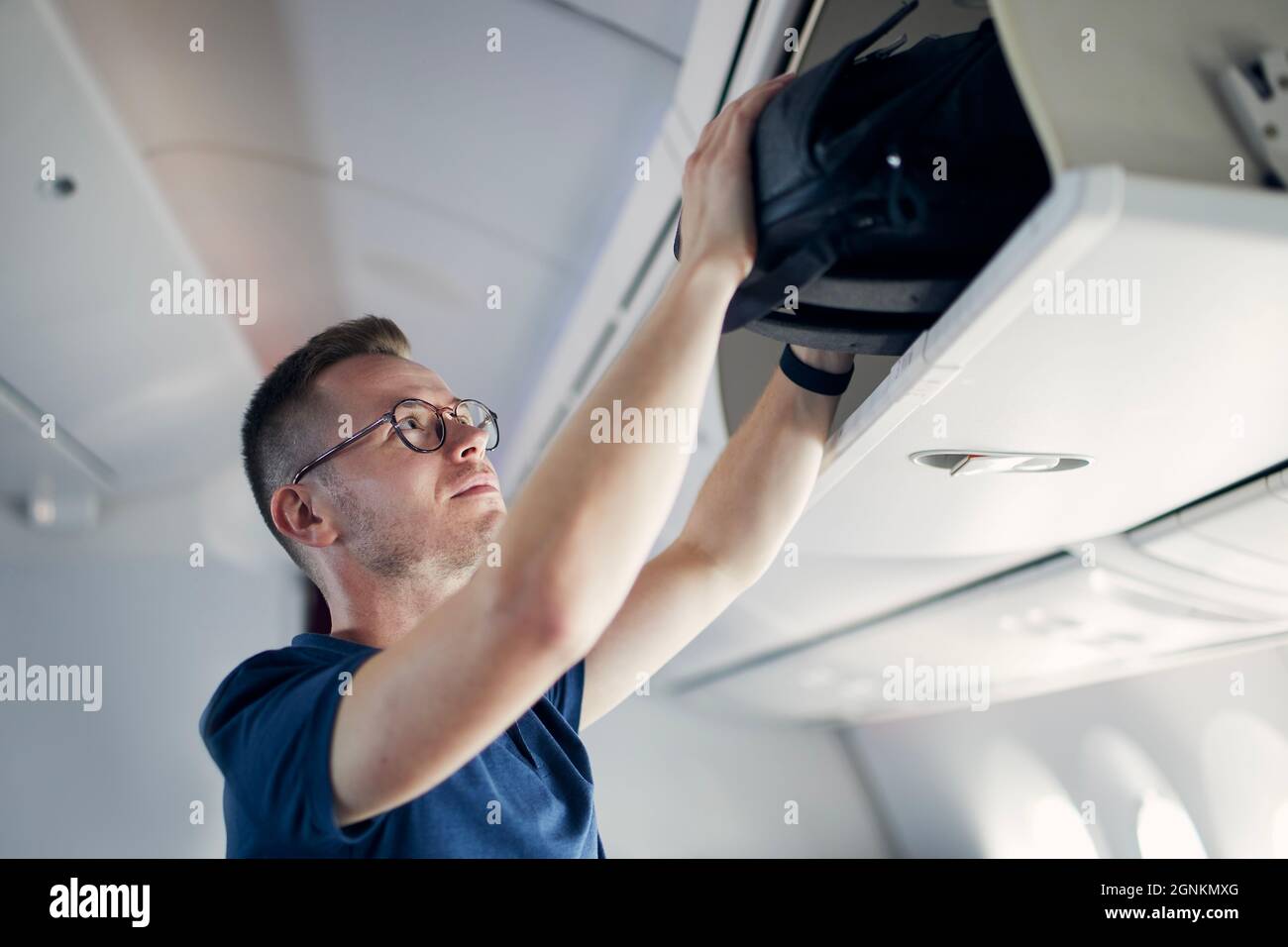 Young man travel by airplane. Passenger putting hand baggage in lockers above seats of plane. Stock Photo
