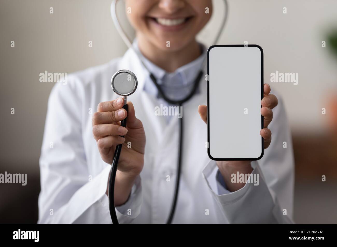 Happy female doctor holding stethoscope, showing blank smartphone screen Stock Photo