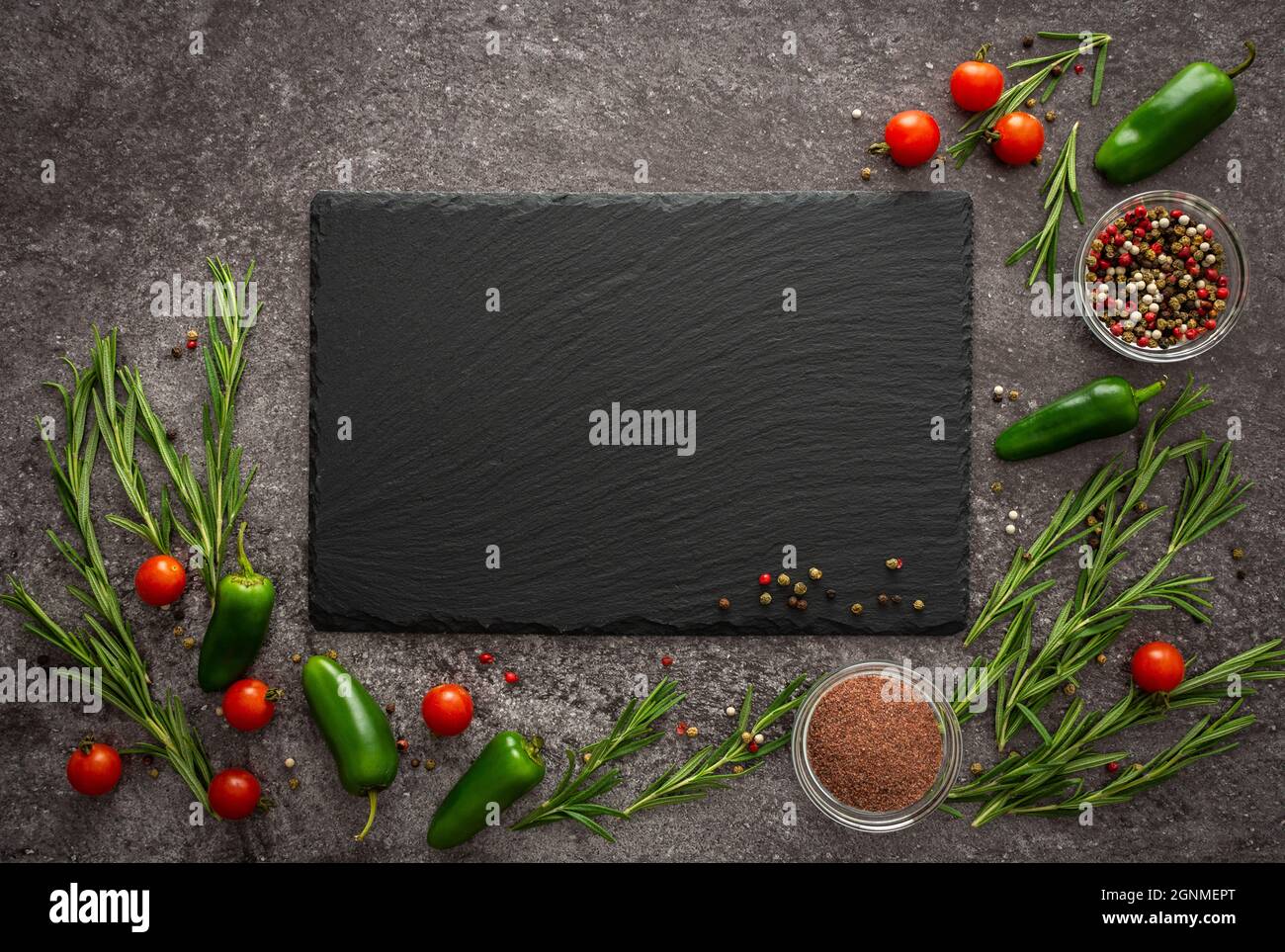 Black empty rectangular slate board on gray background surrounded by fresh cherry tomatoes, jalapeno peppers, spices and rosemary Stock Photo