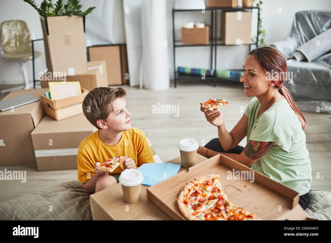 Portrait of young mother and son eating pizza from cardboard box while celebrating moving in to new house, copy space Stock Photo