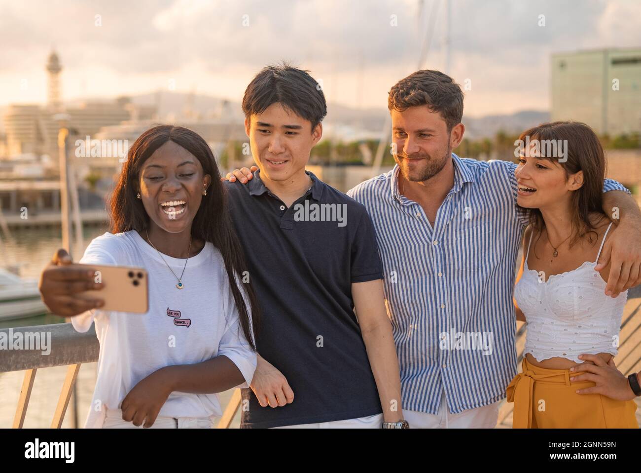 Group of content multiracial young men and women watching funny content on mobile phone or taking selfie while standing together on embankment in summer evening Stock Photo