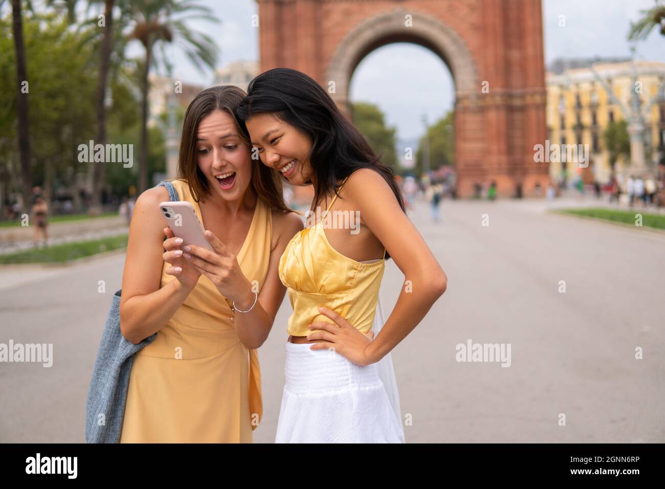 Young multiracial female friends in trendy yellow outfits looking at cellphone screen and laughing while checking photos after sightseeing on street of Barcelona city during summer journey together Stock Photo