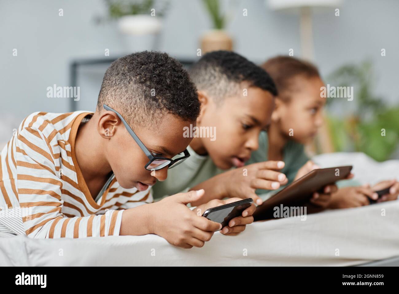Side view portrait of three kids in row using gadgets while lying on bed at home, copy space Stock Photo