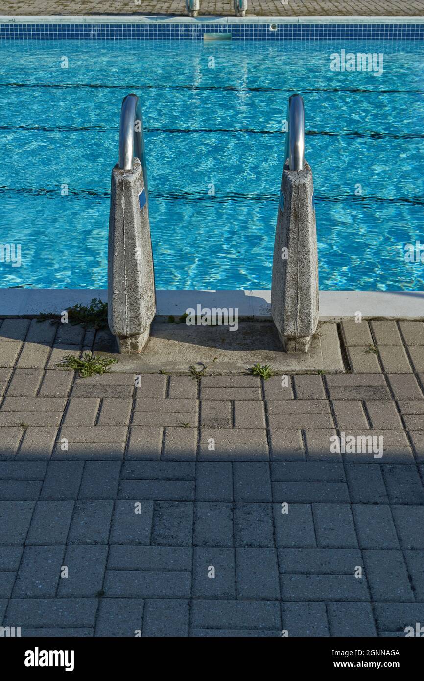 A view from behind a ladder at the edge of an outdoor public pool shows its railings supported by concrete. Stock Photo