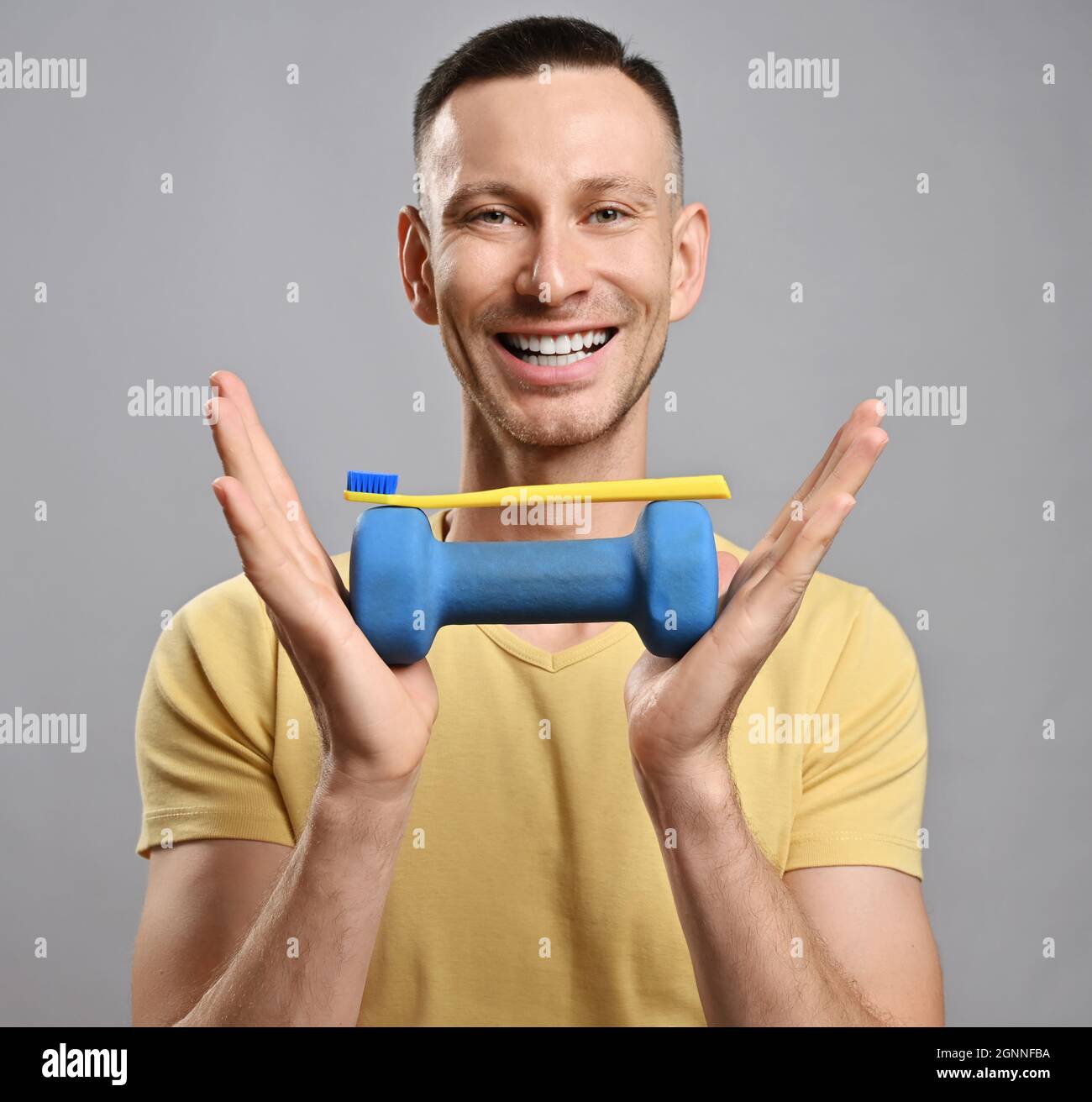 Portrait of happy friendly smiling adult man in yellow t-shirt promoting healthy lifestyle with toothbrush and dumbbell Stock Photo