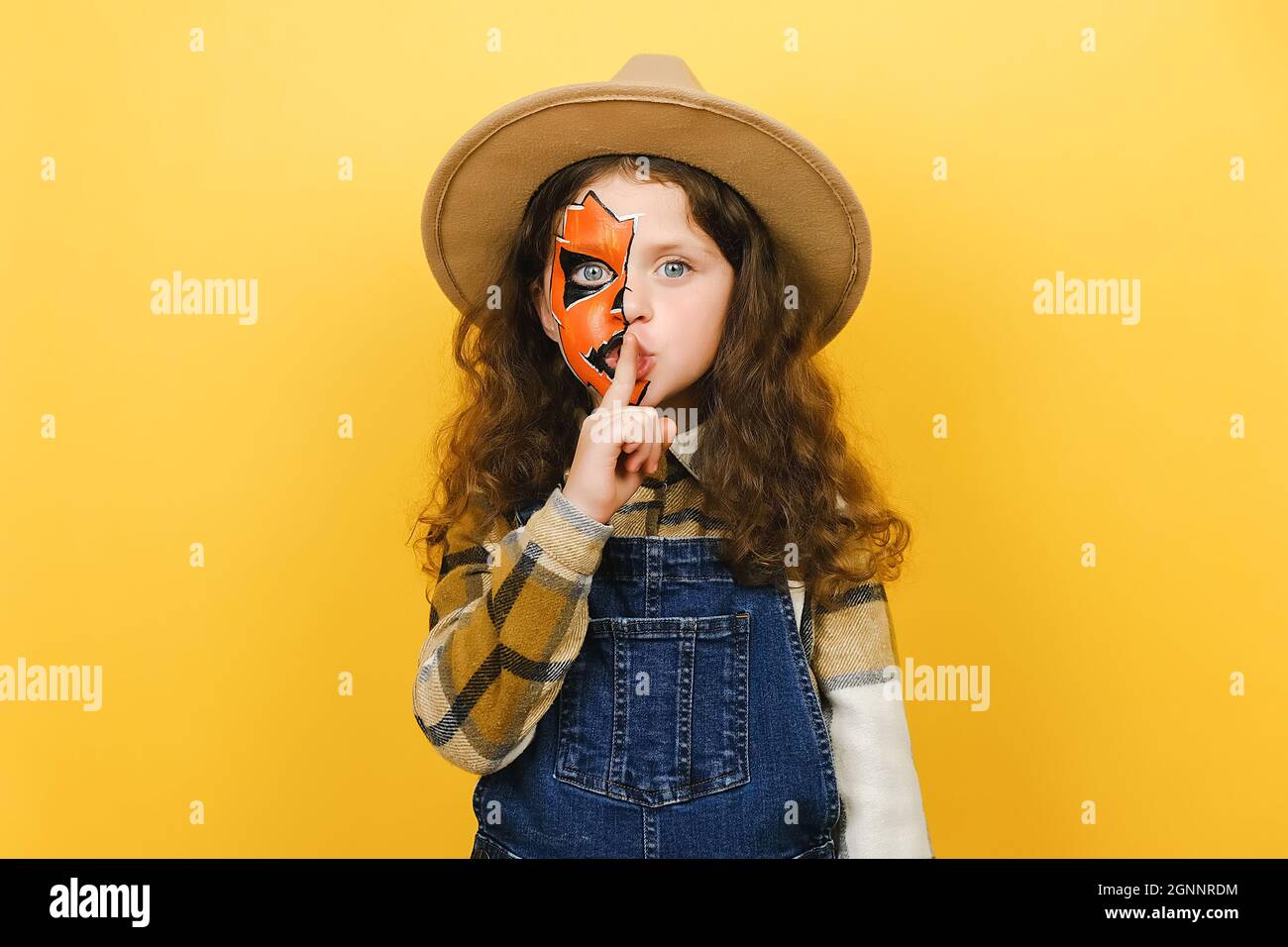 Secret charming fun curly little girl child with Halloween makeup mask wears brown hat, say hush be quiet with finger on lips shhh gesture Stock Photo