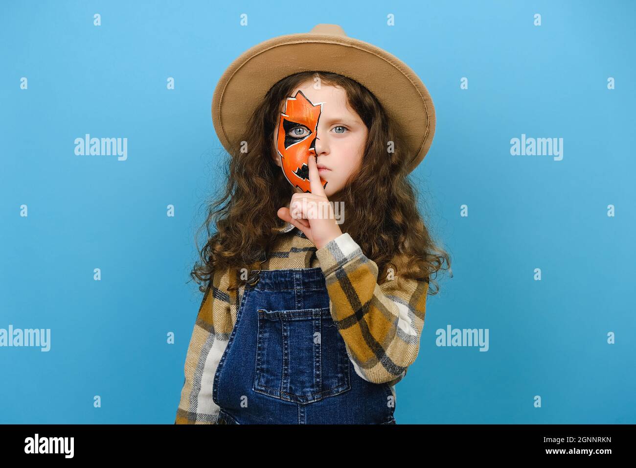 Close up portrait of little girl with Halloween makeup mask wear hat and shirt, say hush be quiet finger on lip shhh gesture isolated on blue wall Stock Photo