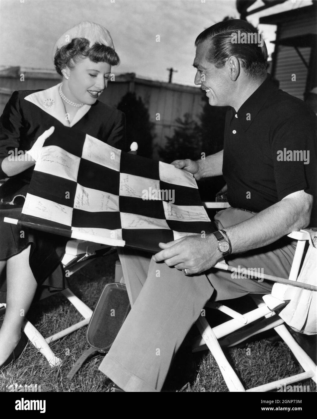 BARBARA STANWYCK shows her Winner's Flag signed to her by the drivers in the  1950 500 mile race at Indianapolis to CLARK GABLE on set location candid  during filming of TO PLEASE