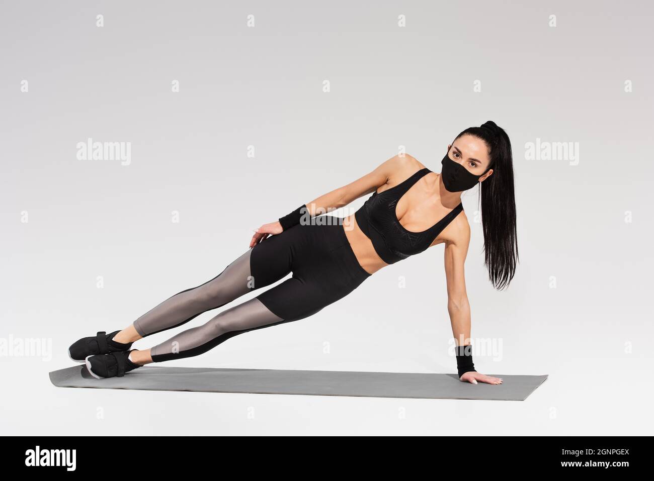 full length of woman in sportswear and black protective mask exercising on fitness mat on grey Stock Photo