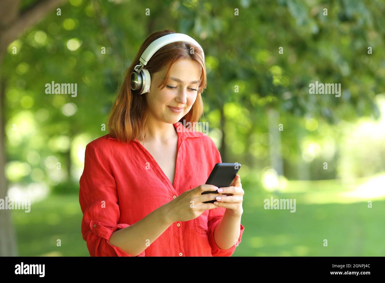 Happy woman in red wearing wireless headphones is waling listening to music on smartphone in a park Stock Photo
