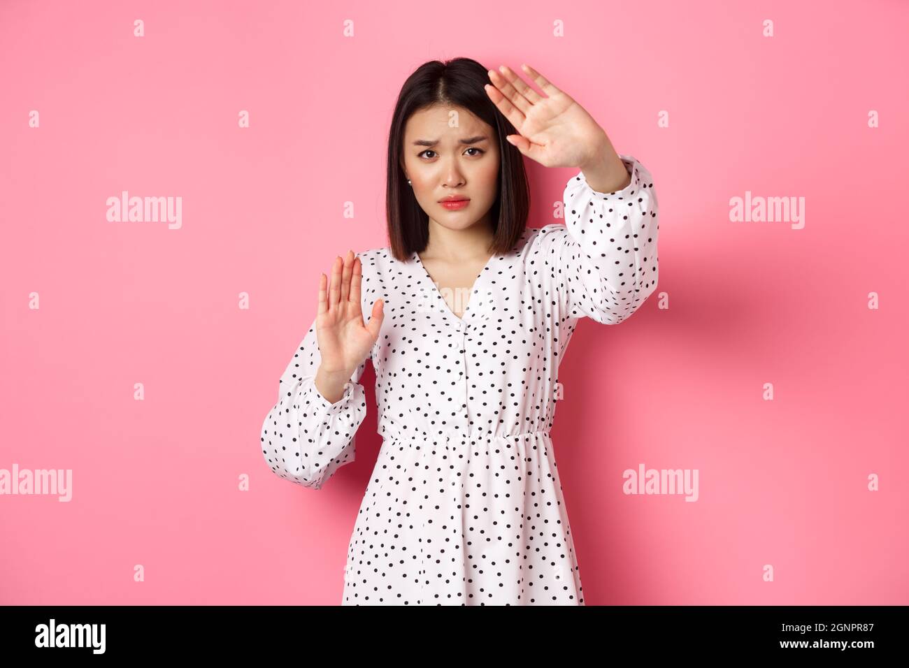 Timid and scared asian woman defending herself, raising arms in protection, victim being attacked, standing over pink background. Stock Photo