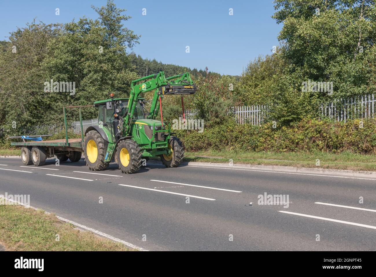 Familiar green John Deere farm tractor on open uphill rural country road, with trailer hitched. Large tyre Deere model is 6115 RC. Stock Photo