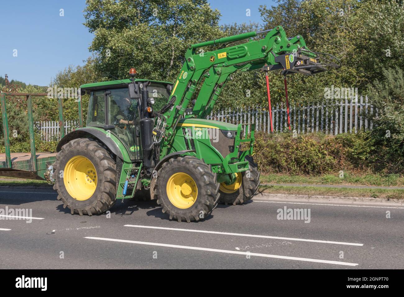 Familiar green John Deere farm tractor on open uphill rural country road, with trailer hitched. Large tyre Deere model is 6115 RC. Stock Photo