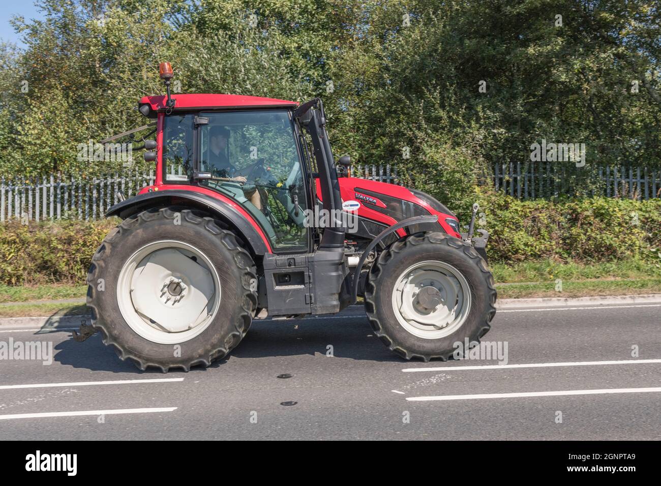 Side profile of red Valtra farm tractor on open uphill rural country road. Valtra is a Finnish tractor brand, this is G Series G136 model. Stock Photo