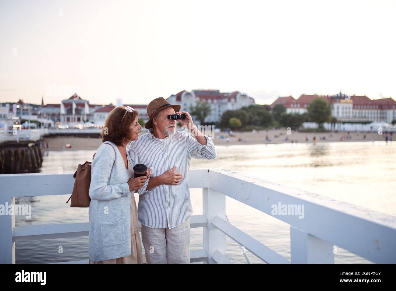 Happy senior couple hugging outdoors on pier by sea, looking at view through binoculars. Stock Photo