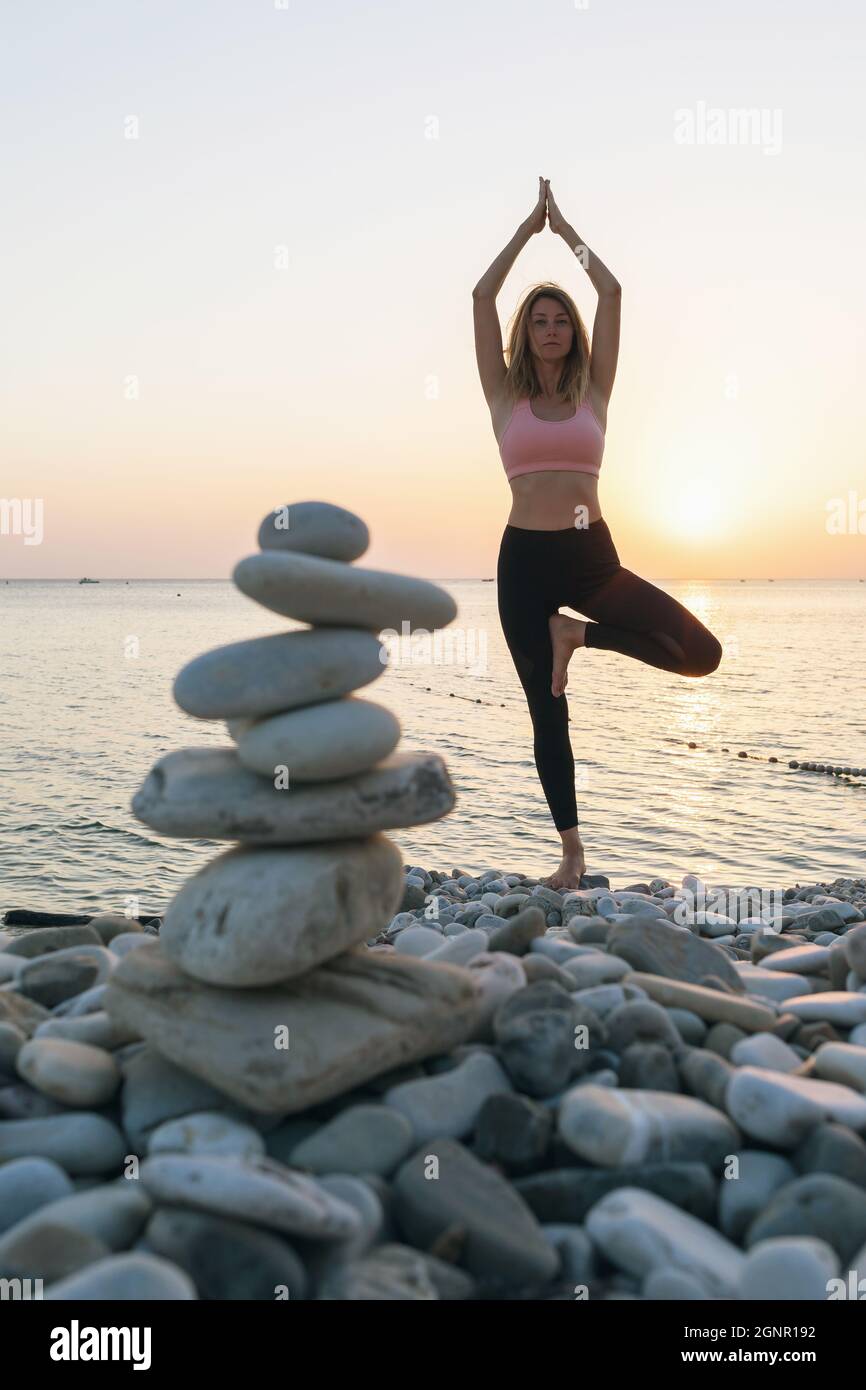 A woman practicing yoga, on the seashore in the evening performs the vrikshasana exercise, a tree pose, a balancing cairn of stones stands nearby in d Stock Photo