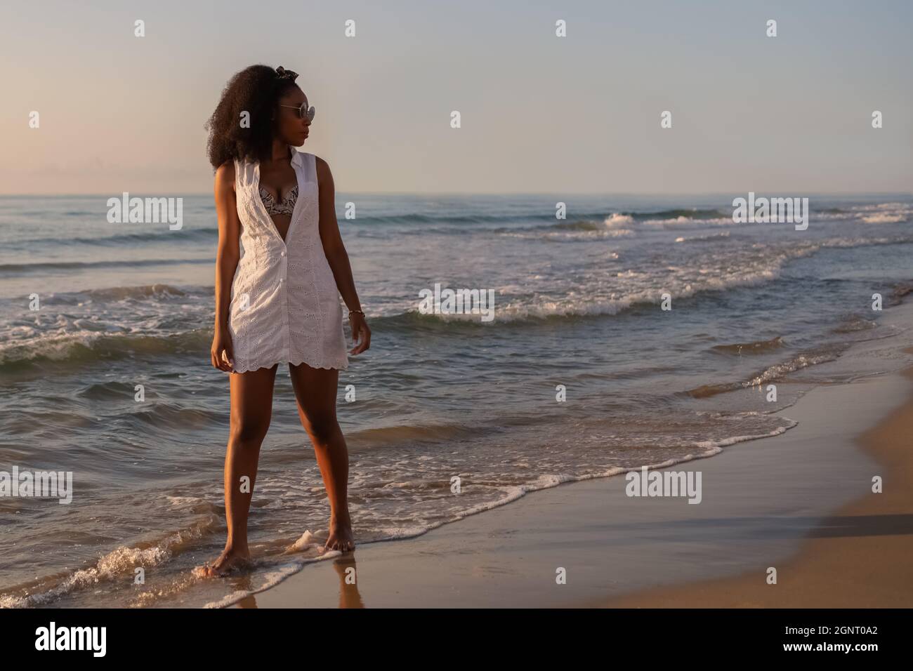 Young adult woman with sunglasses standing along the beach at sunset Stock Photo