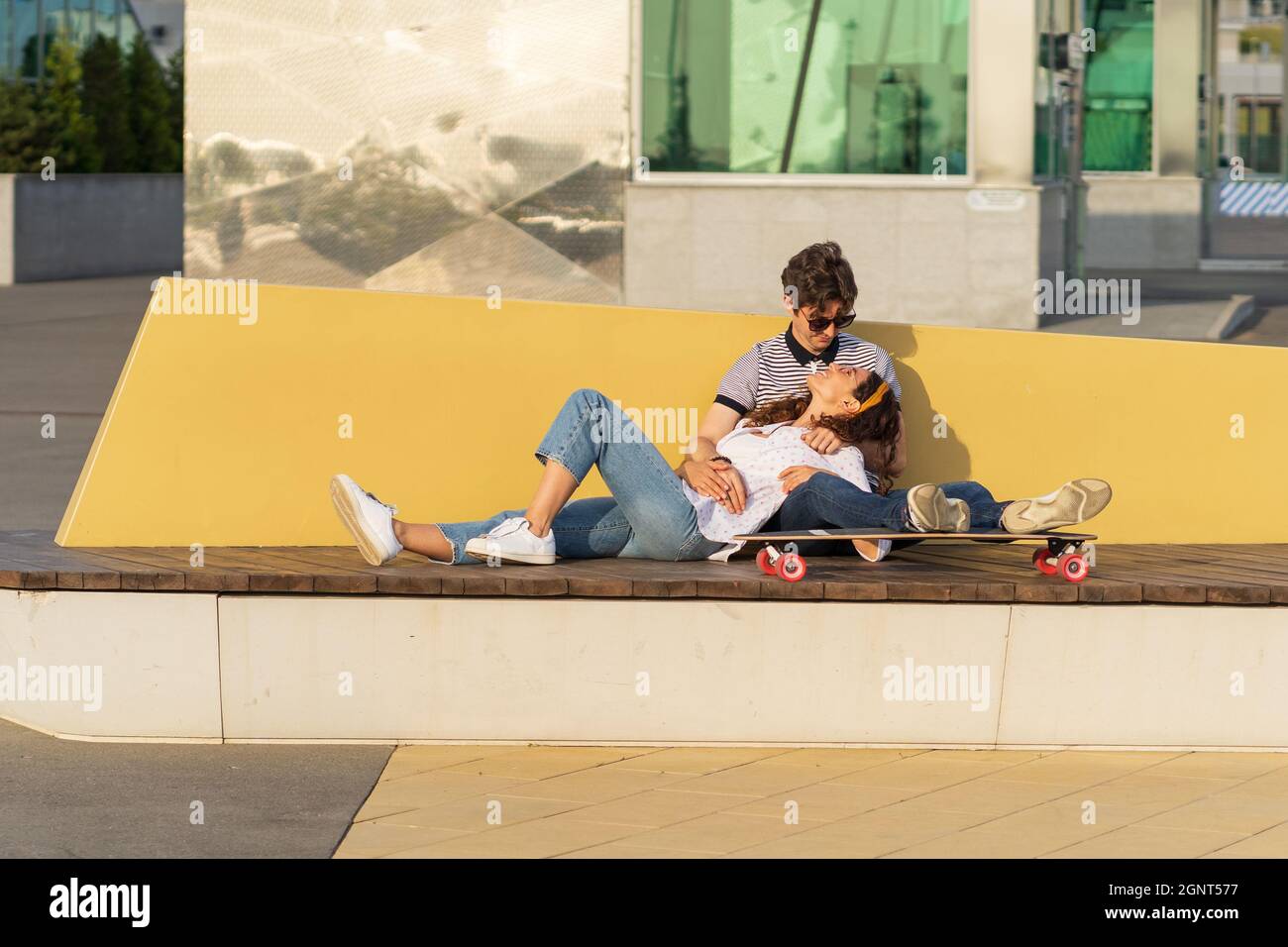 Young trendy couple happy cuddling talking sit on longboard enjoy summer outdoors in urban city park Stock Photo