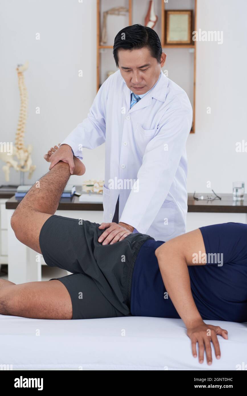 Vertical medium long shot of mature Asian physical therapist wearing white coat working with unrecognizable male patient recovering his hip joints range of motion Stock Photo