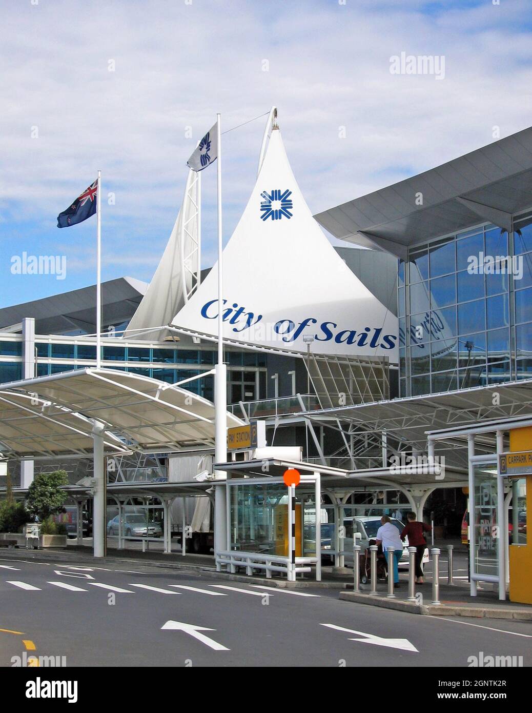 Departures level at Auckland International Airport in Auckland, New Zealand displays the moniker City of Sails outside its main entrance.  The departures level roadway outside the airport entrance is used by travelers pushing luggage during a sunny day in November 2004.  The New Zealand flag waves above the terminal entrance. Stock Photo