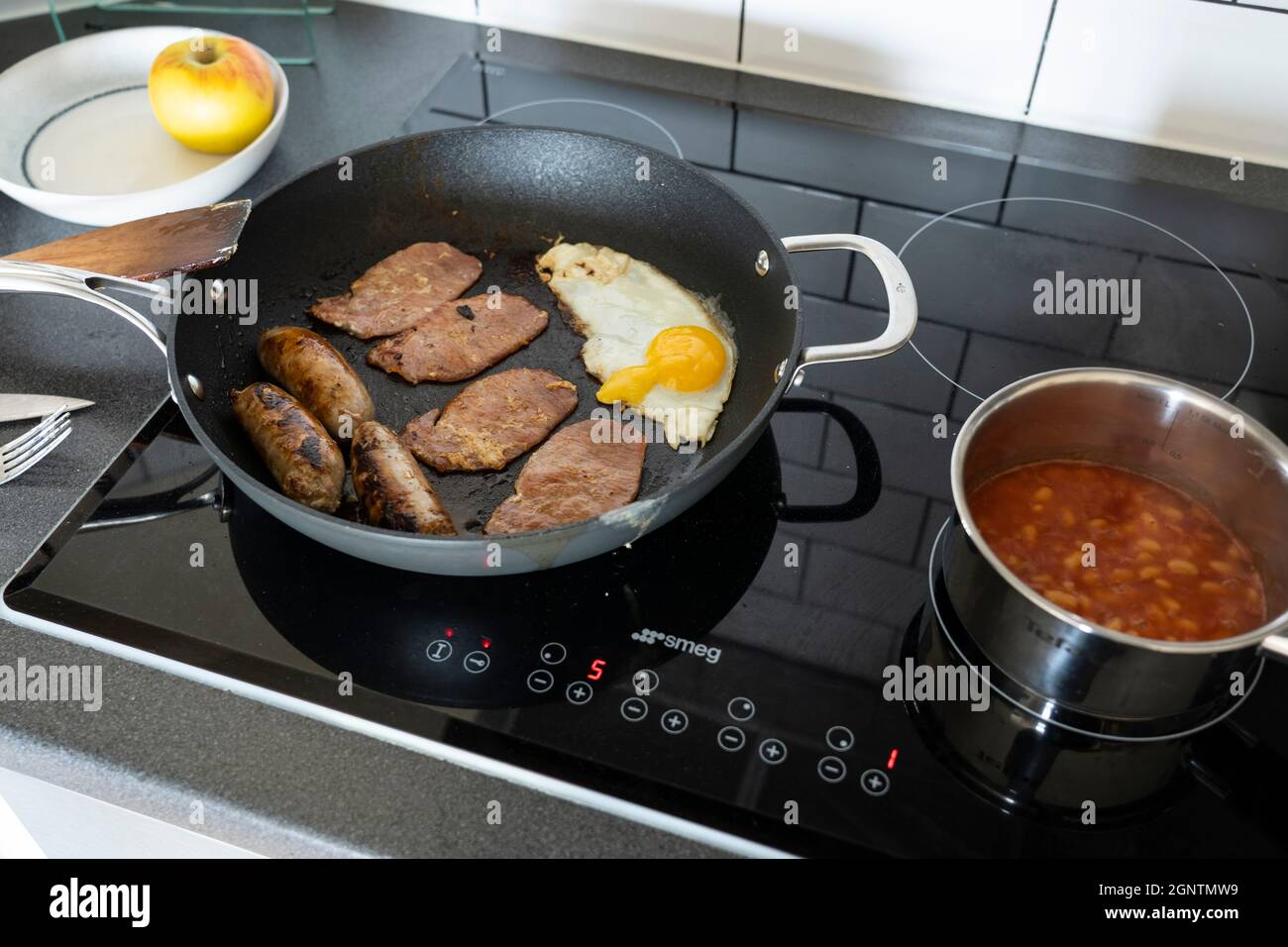 Traditional english breakfast in a kitchen with a fried egg, bacon rashers and sausages cooking in a frying pan, & baked beans in a saucepan. England Stock Photo
