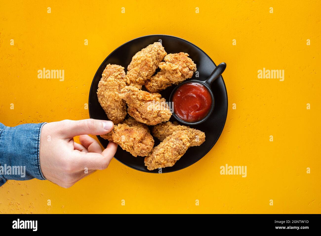 Crispy fried chicken wings with tomato sauce on plate, yellow background. Male hand picking chicken wing, junk food, unhealthy eating concept. Top vie Stock Photo