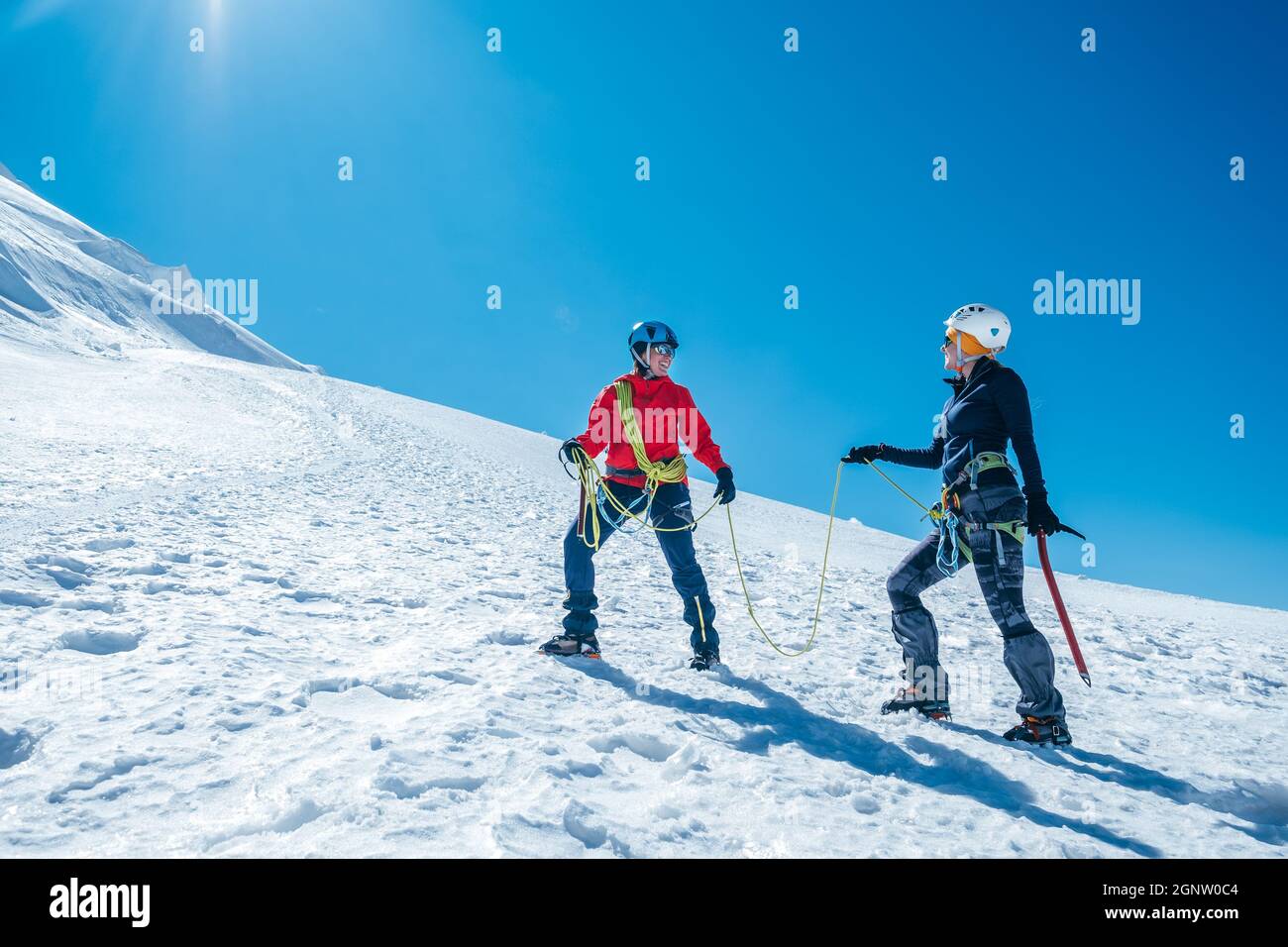 Two laughing to each other young women Rope team ascending Mont blanc du Tacul summit 4248m dressed mountaineering clothes with ice axes on snowy slop Stock Photo