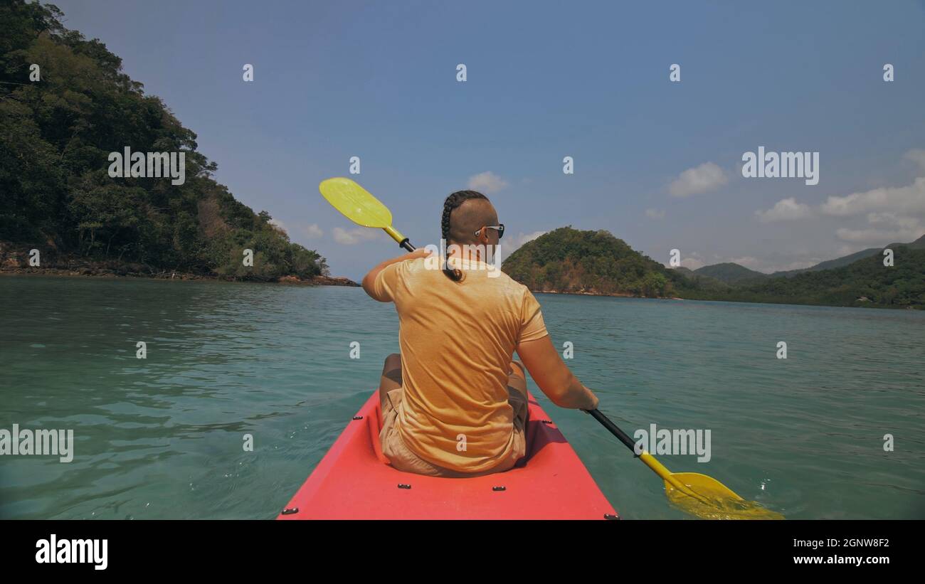 Man with sunglasses and hat rows pink plastic canoe along sea against green hilly islands with wild jungles. Traveling to tropical countries. Stock Photo