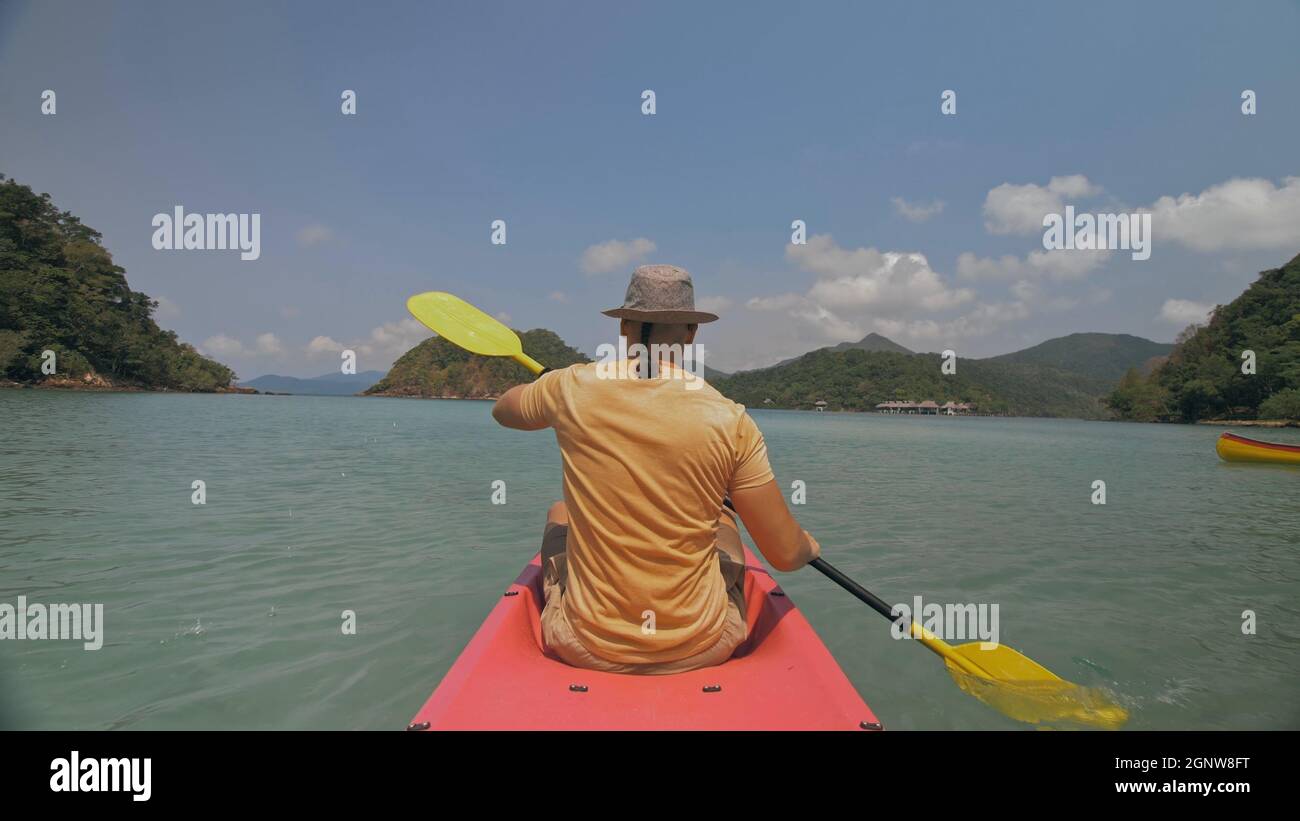 Man with sunglasses and hat rows pink plastic canoe along sea against green hilly islands with wild jungles. Traveling to tropical countries. Stock Photo