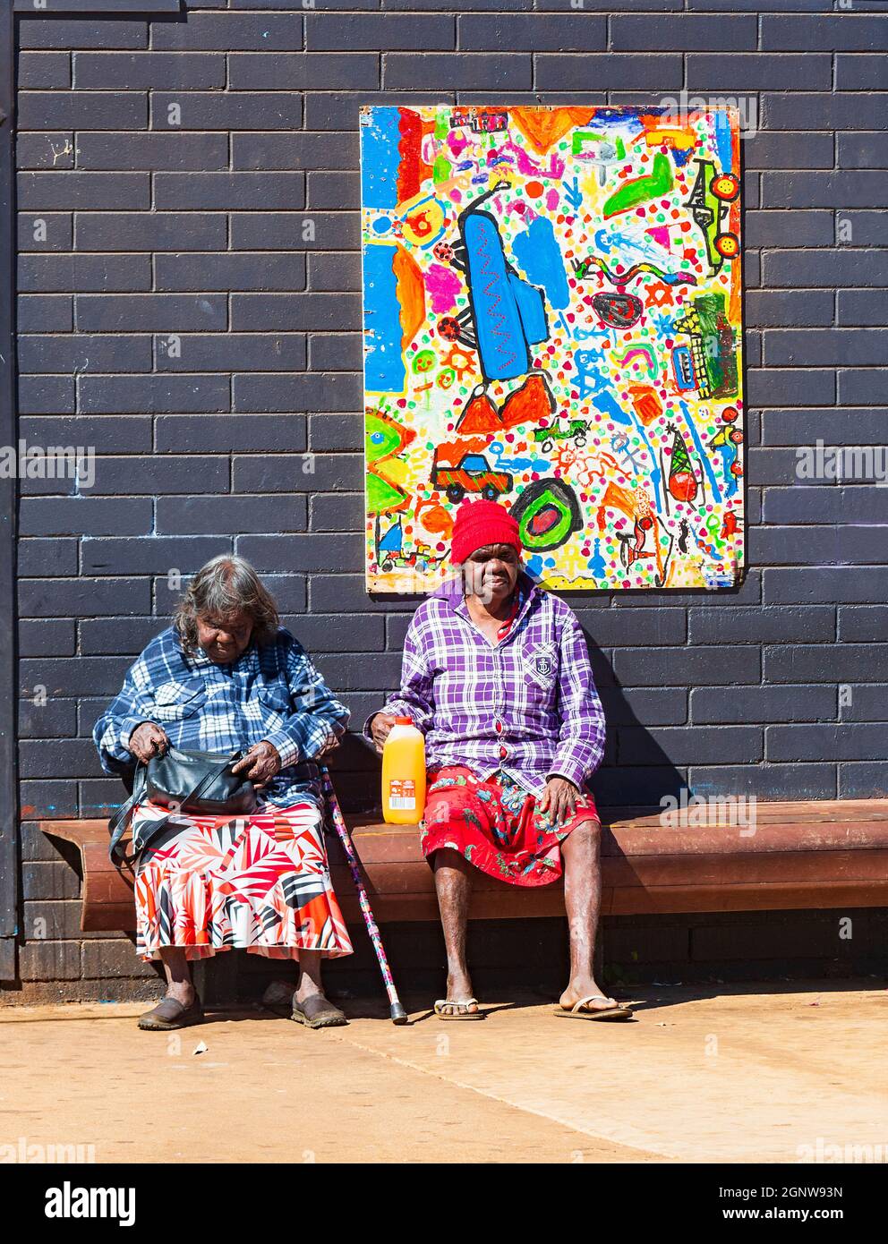 Two elderly Aboriginal women seated on a bench in front of artwork, Halls Creek, Western Australia, Australia Stock Photo