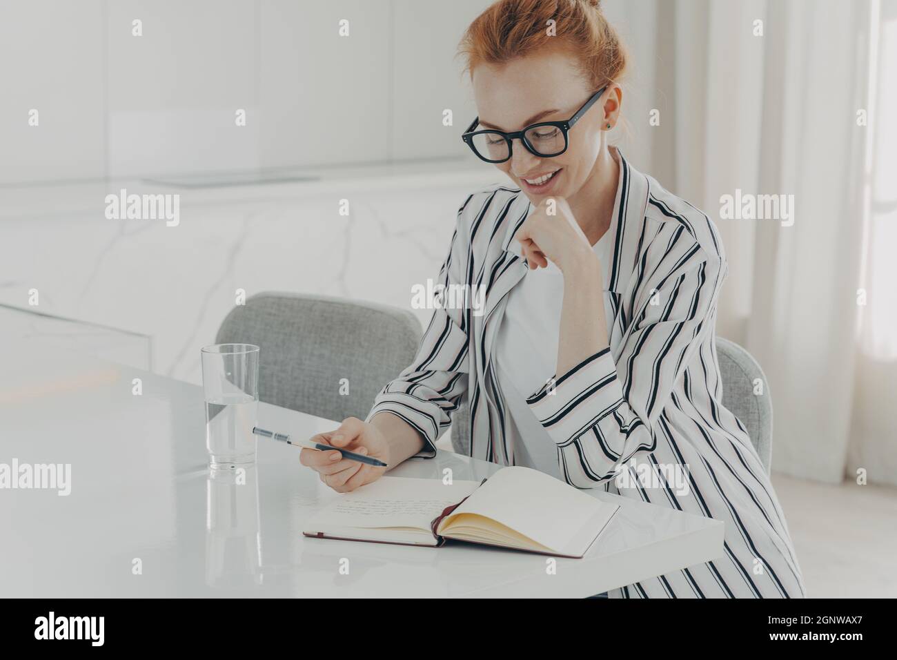 Happy ginger woman taking notes while planning her day in morning at home, drinking pure water Stock Photo